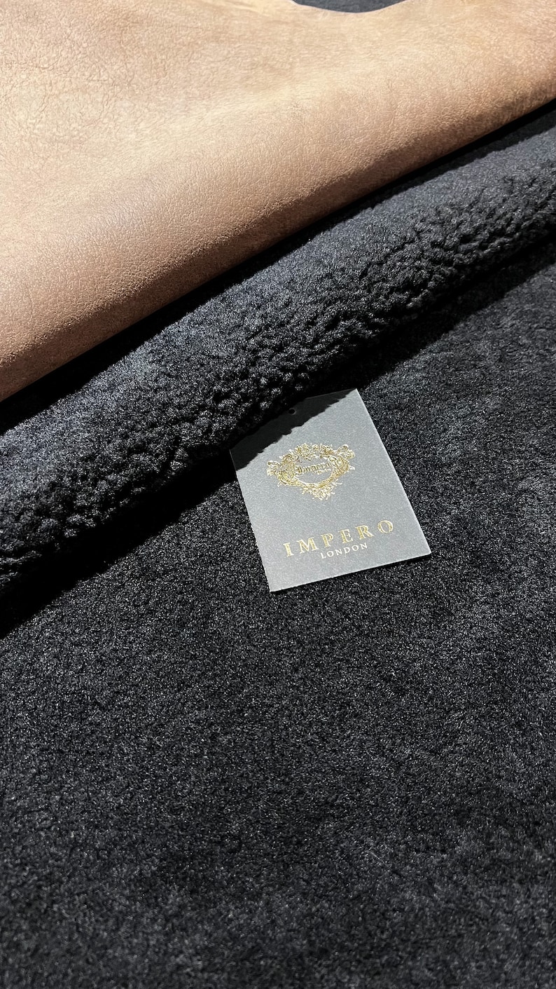 May include: Close-up of a black faux fur fabric with a brown suede fabric folded over it. A white tag with gold lettering reads "Impero London".