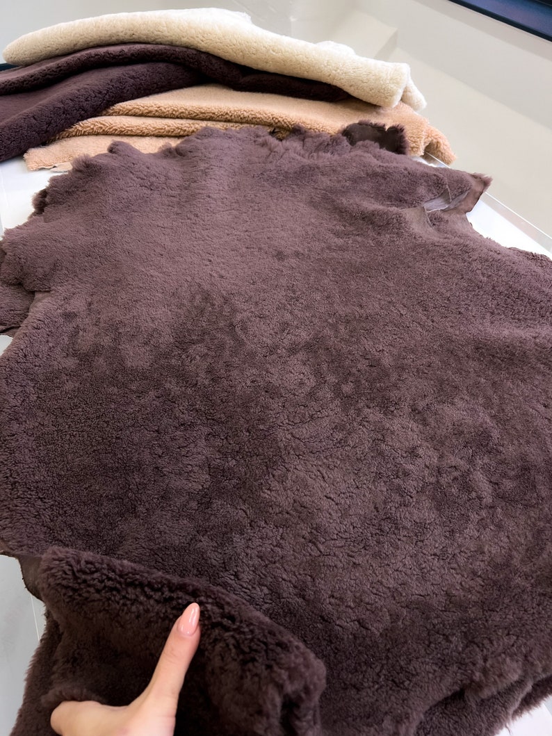 May include: Close-up of several pieces of faux fur fabric in various shades of brown and beige. The fabrics have a soft, textured appearance, with a deep, rich brown piece in the foreground.