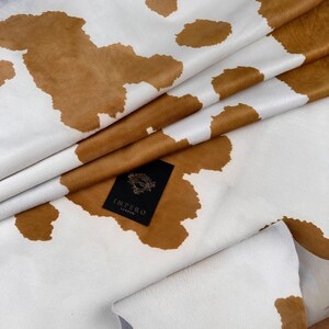 May include: Close-up of a cowhide leather with a white background and brown patches. The leather is folded, revealing its texture and natural patterns. A black tag with the text "IMPERO LONDON" is visible.