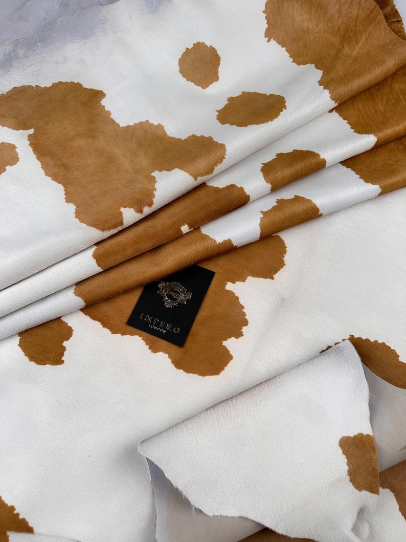 May include: Close-up of a cowhide leather with a white background and brown patches. The leather is folded, revealing its texture and natural patterns. A black tag with the text "IMPERO LONDON" is visible.