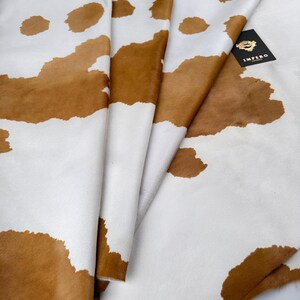 May include: A folded cowhide rug with a brown and white spotted pattern. The hide has a soft texture and a natural appearance. A black tag with gold text is visible.