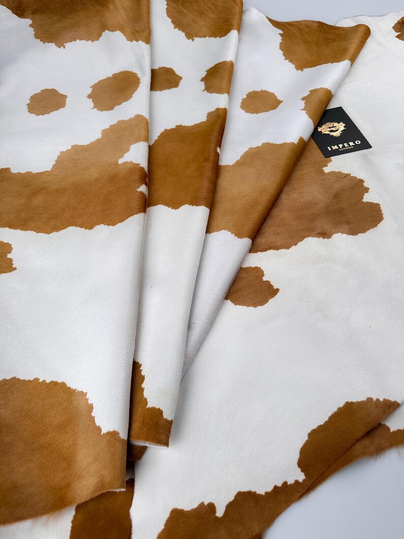 May include: A folded cowhide rug with a brown and white spotted pattern. The hide has a soft texture and a natural appearance. A black tag with gold text is visible.