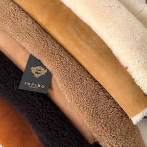 May include: A stack of five different colored sheepskin hides. The hides are arranged in a graduated color scheme from white to brown. The top hide is white, the second is a light beige, the third is a light brown, the fourth is a medium brown, and the fifth is a dark brown. The hides are all soft and fluffy. The label on the bottom hide reads "Impero London".