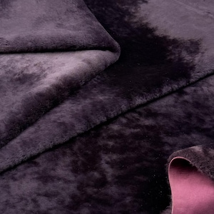 Könnte beinhalten: Nahaufnahme eines flauschigen Kunstfellstoffs in tiefen Lila- und Schwarztönen. Der Stoff ist gefaltet und zeigt eine kontrastierende rosa Unterseite. Die Textur wirkt weich und luxuriös, geeignet für verschiedene Bastelprojekte.
