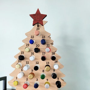 May include: Wooden Christmas tree advent calendar with 24 numbered slots for small bottles. The tree is decorated with a red star on top and has the text "Making Spirits Bright" on the star. The bottles are filled with various colored liquids.