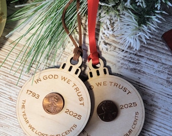 Pode incluir: Duas decorações de Natal de madeira com uma moeda de um centavo no centro. Os ornamentos têm o texto "IN GOD WE TRUST", "THE YEAR WE LOST OUR CENTS" e os anos 1793 e 2025. Um tem uma corda de camurça castanha e o outro uma fita vermelha.