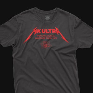 May include: Dark gray t-shirt with a red graphic that reads "MK ULTRA PSYCHOLOGICAL WARFARE TOUR 1973". The graphic includes a stylized skull and a logo. The shirt has a crew neck and short sleeves.
