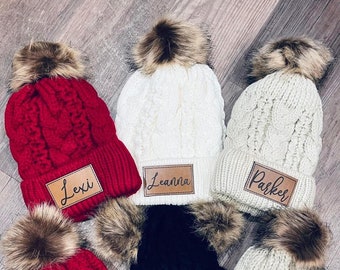 May include: Six knitted winter hats with faux fur pom poms and personalized leather patches. The hats are in red, white, and black. The names on the patches are Lexi, Leanna, Parker, Payton, Kingsley, and Brooklyn.