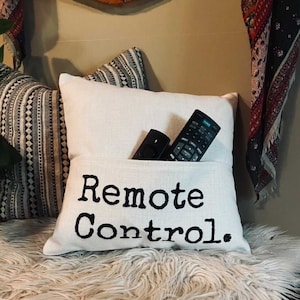 May include: A white linen pillow with a pocket, embroidered with the words "Remote Control" in black. Two black remote controls are in the pocket. The pillow is on a fluffy white surface.