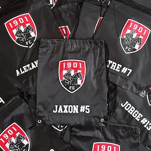 May include: Black drawstring bags with a red and white emblem featuring a rider on a horse, the text "1901 FC", and personalized names and numbers in white. The bags are made of a shiny material.