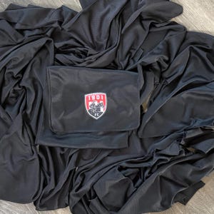 May include: A black fabric item with a textured surface featuring a shield-shaped patch in the center. The patch displays the text 1901 FC above a graphic of a horse and rider in white, black, and red colors against a dark background.