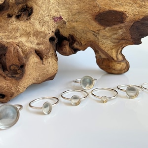 May include: Six silver rings with clear glass or resin insets. The rings are arranged in a row on a white surface.