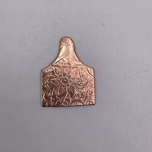 May include: A copper-colored metal tag with a floral design. The tag has a pointed top and a rounded bottom.