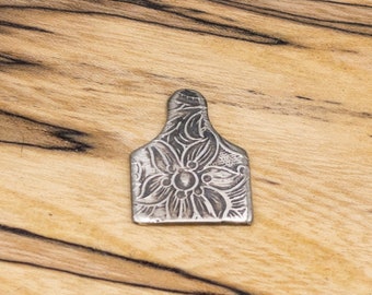 Op de afbeelding: Een zilveren bedel in de vorm van een oorstempel voor vee met een bloemenmotief.