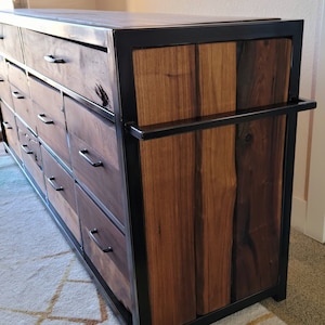 May include: A dark wood dresser with a black metal frame and multiple drawers. The dresser has a unique design with a single large drawer on the top and smaller drawers below. The dresser is sitting on a white rug with a geometric pattern.