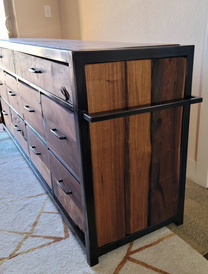 May include: A dark wood dresser with a black metal frame and multiple drawers. The dresser has a unique design with a single large drawer on the top and smaller drawers below. The dresser is sitting on a white rug with a geometric pattern.