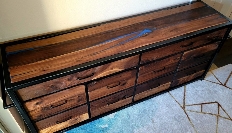 May include: A dresser with a dark wood finish and a black metal frame. The top of the dresser has a blue resin inlay. The dresser has eight drawers with black metal handles.
