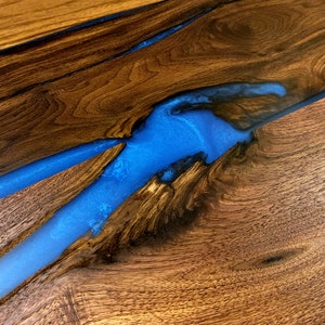 May include: A close-up of a wooden table top with a blue epoxy resin river running through it. The wood has a rich brown color and the resin is clear and shiny.
