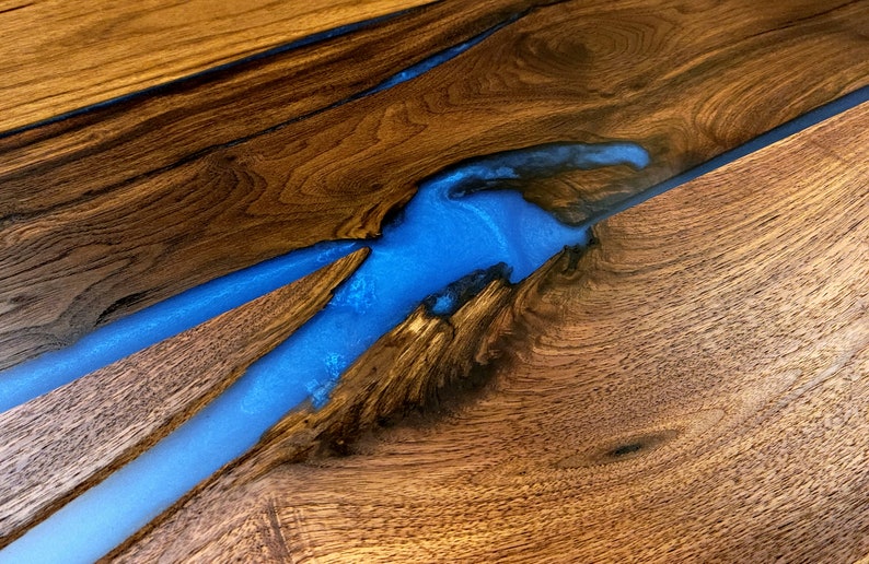 May include: A close-up of a wooden table top with a blue epoxy resin river running through it. The wood has a rich brown color and the resin is clear and shiny.