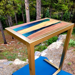 May include: A gold metal side table with a wooden top and a green and blue epoxy resin inlay. The table has a simple, modern design and would be a great addition to any home.