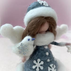May include: A handcrafted felt angel figurine in a winter theme. The angel wears a blue dress with snowflake accents, a white fur-trimmed coat, and a matching hat. It holds a white bird with a snowflake design and has white wings. The angel has brown hair.