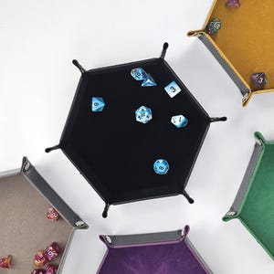May include: A set of six hexagonal dice trays in various colors, including black, brown, green, purple, and yellow. Each tray has a felt lining and is designed to hold dice for tabletop games.