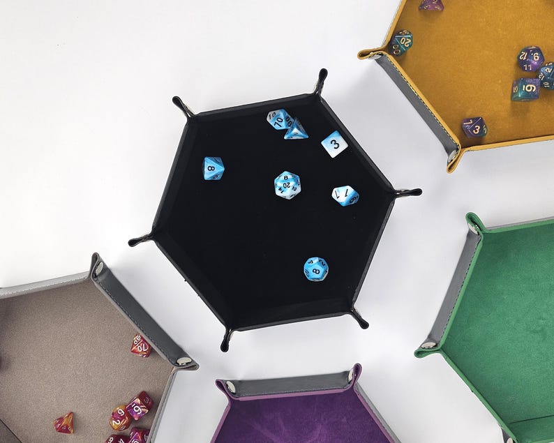 May include: A set of six hexagonal dice trays in various colors, including black, brown, green, purple, and yellow. Each tray has a felt lining and is designed to hold dice for tabletop games.