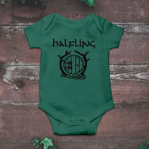 Puede incluir: Un body de bebé verde con la palabra "halfling" en escritura negra sobre una ilustración negra de una puerta redonda. La puerta está enmarcada por ramas de árboles y descansa sobre un lecho de piedras.