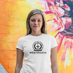 Peut inclure: Un t-shirt blanc avec un logo noir représentant une couronne, une couronne de laurier et une croix. Le texte "Correctional Services" et "Wentworth" est imprimé sous le logo.