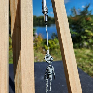 Può includere: Una scultura in filo metallico di una persona appesa a una clip argentata. La scultura è realizzata in filo metallico grigio ed è appesa a un cavalletto di legno.
