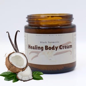 Op de afbeelding: Een bruine glazen pot Black Turmeric Healing Body Cream met een wit etiket. De afbeelding bevat een gehalveerde kokosnoot, vanillestokjes en groene bladeren, wat op natuurlijke ingrediënten wijst.