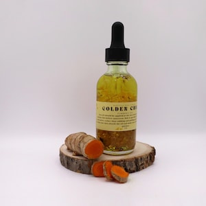 May include: A glass bottle with a dropper filled with a golden liquid and labelled "Golden Chic Turmeric Oil". The bottle is sitting on a wooden slice with pieces of turmeric root.