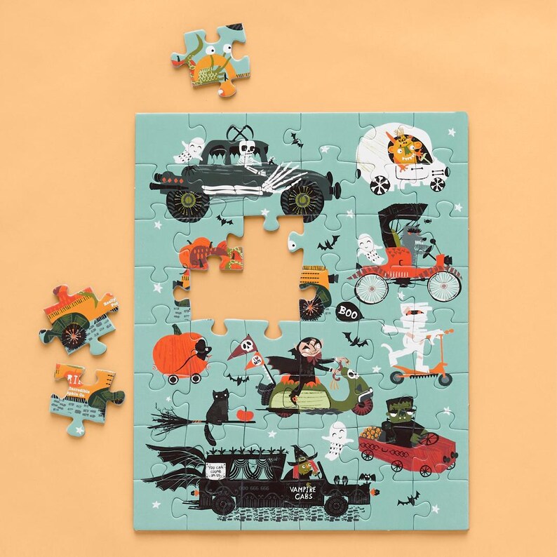 Op de afbeelding: Een kleurrijke Halloween-puzzel met een blauwe achtergrond en verschillende griezelige voertuigen, waaronder een auto in de vorm van een pompoen, een monstertruck en een vampierauto. De puzzel is gedeeltelijk voltooid, met enkele ontbrekende stukken.