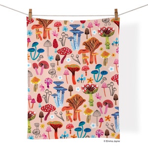 May include: A pink tea towel with a colorful mushroom pattern. The mushrooms are in various shapes and sizes, with red, orange, yellow, blue, and green accents. There are also small white flowers scattered throughout the design.