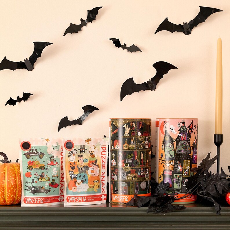 Op de afbeelding: Drie Halloween-puzzels in cilindrische dozen met zwarte vleermuizen op een beige muur. De puzzels zijn getiteld "Puzzle Snax" en bevatten griezelige illustraties.