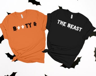 Puede incluir: Dos camisetas con temática de Halloween. Una camiseta naranja con el texto "Boo-ty &" en negro con dos fantasmas blancos. La otra camiseta negra con el texto "The Beast" en blanco con letras que gotean.