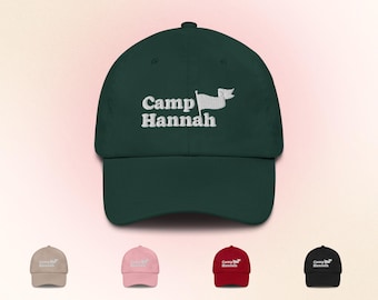 Puede incluir: Una gorra de béisbol verde con bordado blanco que dice "Camp Hannah" con un diseño de bandera blanca. La gorra está disponible en otros colores.