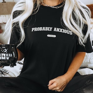 May include: Black t-shirt with the words "PROBABLY ANXIOUS" in white, and a small rectangular graphic below. A black cauldron-shaped mug with "WITCHES BREW" is held in the hand. The mug has a handle and three small feet.
