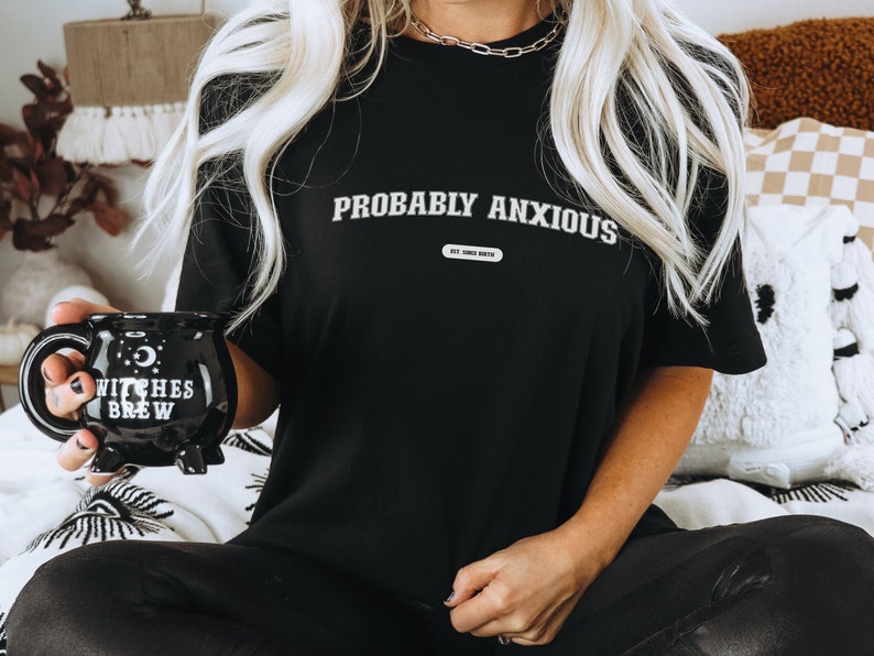 May include: Black t-shirt with the words "PROBABLY ANXIOUS" in white, and a small rectangular graphic below. A black cauldron-shaped mug with "WITCHES BREW" is held in the hand. The mug has a handle and three small feet.