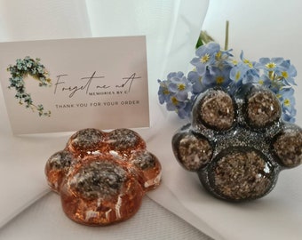 May include: Two resin paw print ornaments, one in a copper color and one in a gray color. The ornaments are decorated with glitter and have a textured surface. The ornaments are sitting on a white surface. A card with the text "Forget me not, Memories by C, Thank you for your order" is in the background.
