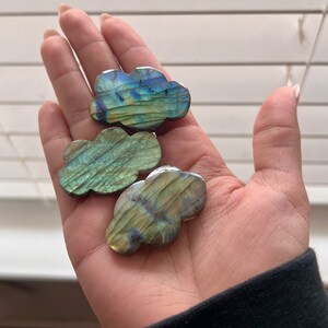 May include: Three iridescent labradorite crystal cloud shapes in a hand. The crystals are green, blue, and gold.