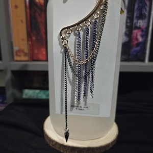 May include: A gold-toned ear cuff featuring multiple dangling chains in black, silver, and blue. The piece includes a small black spike charm at the end of the longest chain. A white display card behind the jewelry features the text Attack on Titan.