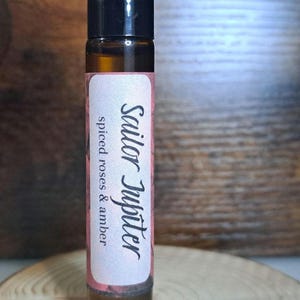 May include: A brown glass roller bottle with a black cap. The label is pink and says "Sailor Jupiter" and "spiced roses & amber".