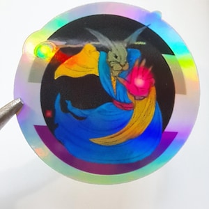 May include: Holographic sticker featuring a fantasy character with a rabbit-like head, wearing a blue robe and a yellow cape. The character's hand glows pink, set against a black background within a circular design with rainbow edges.