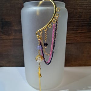 May include: A gold-colored ear cuff with dangling chains and decorative beads. The ear cuff features a curved design that wraps around the ear, with a small sword pendant hanging from the bottom. The chains are pink, black, and gold.