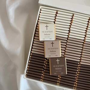 Peut inclure: Une boîte de barres chocolatées emballées individuellement avec des accents dorés. Chaque barre a une petite étiquette carrée avec une croix, le texte "MY BAPTISM", un nom et une date.
