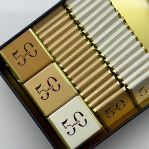 Könnte beinhalten: Eine schwarze rechteckige Schachtel gefüllt mit einzeln verpackten Pralinen. Die Pralinen sind in goldenen und weißen Verpackungen. Einige Pralinen tragen die Zahl "50" und den Namen "Renata".
