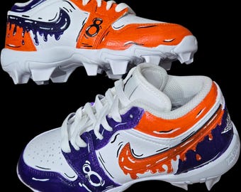 May include: White and orange baseball cleats with purple accents and the number 8. The cleats feature a swoosh design and drip-style paint details. The cleats have white laces and a cleated sole.