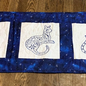 Op de afbeelding: Een blauwe en witte gestikte tafelloper met drie geborduurde katten. Elke kat zit en heeft een paisley-ontwerp.