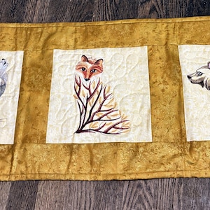 May include: A golden-yellow quilted table runner featuring three embroidered animal designs. The designs include a howling wolf, a fox, and a bear, each set within a cream-colored square. The runner is approximately 40 inches long.